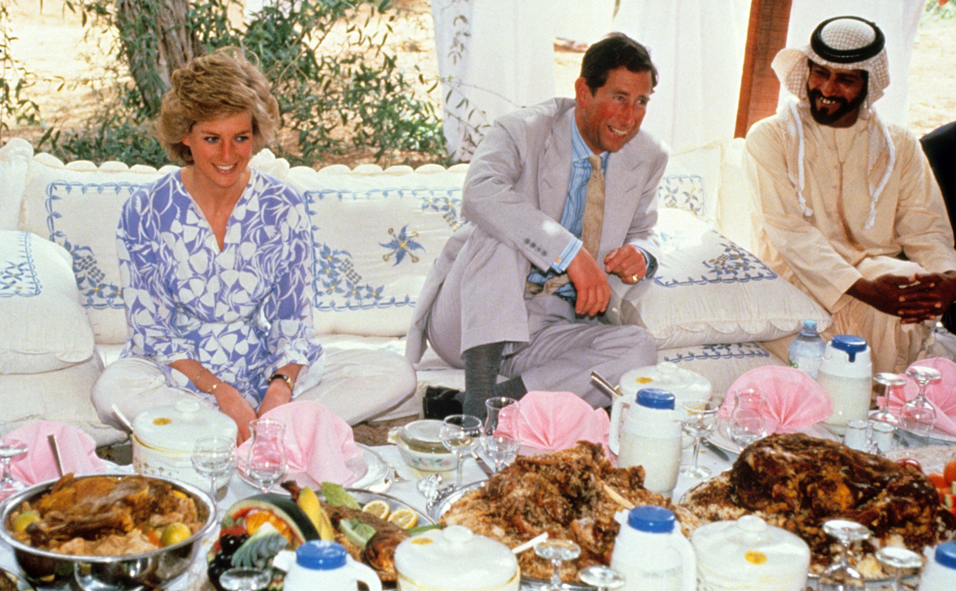 Princess Diana sitting on a cushion next to Prince Charles in front of a big table of food