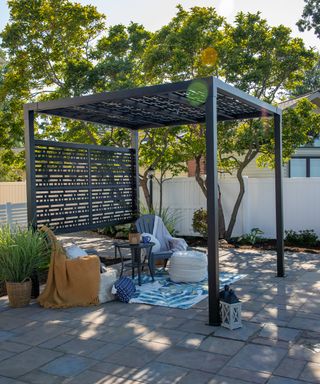 A black metal pergola with slatted screens on a patio blue outdoor rug, gray Adirondack chairs and cozy soft furnishings in yellow, blue and cream