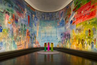 View of the Musée d’Art Moderne de Paris tribute to Yves Saint Laurent featuring three mannequins displaying colourful fashion pieces in a room with curved colourful walls and dark coloured flooring