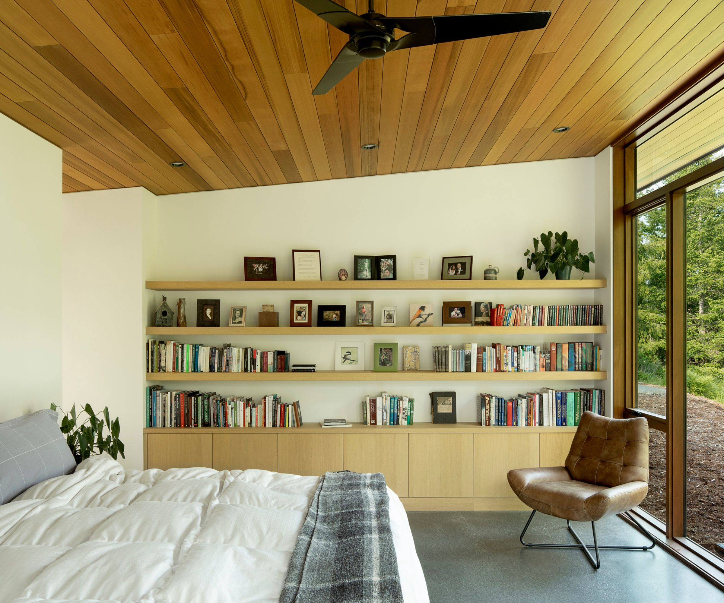 Midcentury modern bedroom with leather armchair, wooden open shelving decorated with books and pictures, and bed with white bedding, and glass doors onlooking the garden