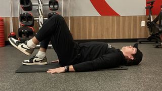 Man lies on his back on exercise mat with knees bent, one foot flat on the floor and the other raised. He wears Nike trainers, black pants, black sweatshirt and black cap.