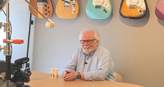 Guitar guru Trevor Wilkinson sits at a table with guitars hanging on the wall behind him