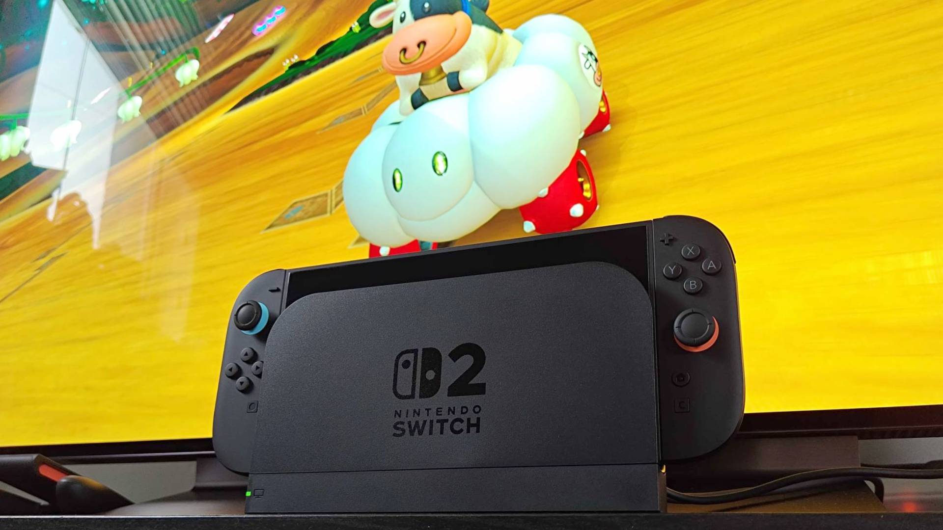 Nintendo Switch 2 sitting in dock with LG OLED TV in backdrop and Mario Kart World gameplay on screen featuring Cow in cloud car.