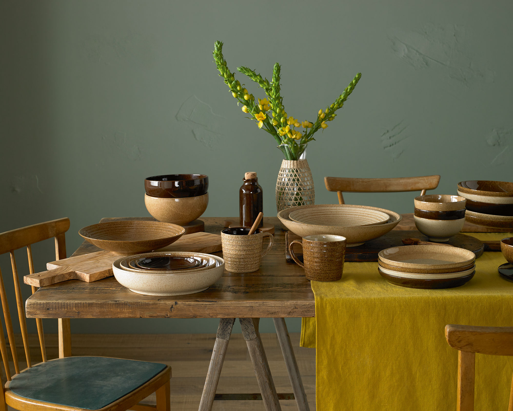 A rustic wooden dining table is set with various stoneware dishes from the Denby Studio Craft collection, featuring earthy tones of chestnut, birch, and walnut. The arrangement includes bowls, plates, and mugs with speckled and glazed finishes. A glass vase wrapped in woven seagrass holds bright yellow flowers in the center. The table is draped with a mustard yellow linen runner, and the scene is set against a muted sage green textured wall. Two wooden spindle-back chairs are partially visible, one with a teal seat cushion.