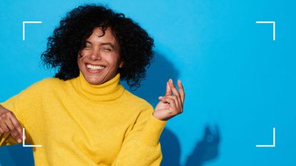 Woman clicking her fingers and laughing, wearing a bright jumper against a plain wall, representing sex positivity