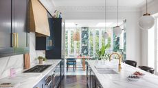 Bright kitchen and dining area with marble-esque worktops, dark navy cabinets, a goldbronze range hood, and low-hanging lights. Kitchen has a wooden floor and the dining area, in background, has a blue-and-white tiled floor with floral patterned wallpaper.