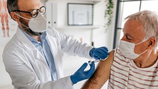 a male doctor wearing a white N95 mask gives a vaccine to an older male patient wearing a white surgical mask