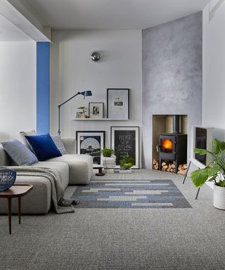 A grey living room with grey and blue carpet tiles, blue floor lamp, framed wall art on floor and floating shelves, a traditional black woodburning stove in concrete alcove and potted houseplant