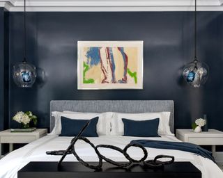 Blue painted bedroom with abstract artwork above bed, two low hanging round glass pendants above white bedside tables, light blue upholstered headboard, white linen, blue cushions and throw, black cabinet at end of bed, sculptural black metal artwork positioned on top.