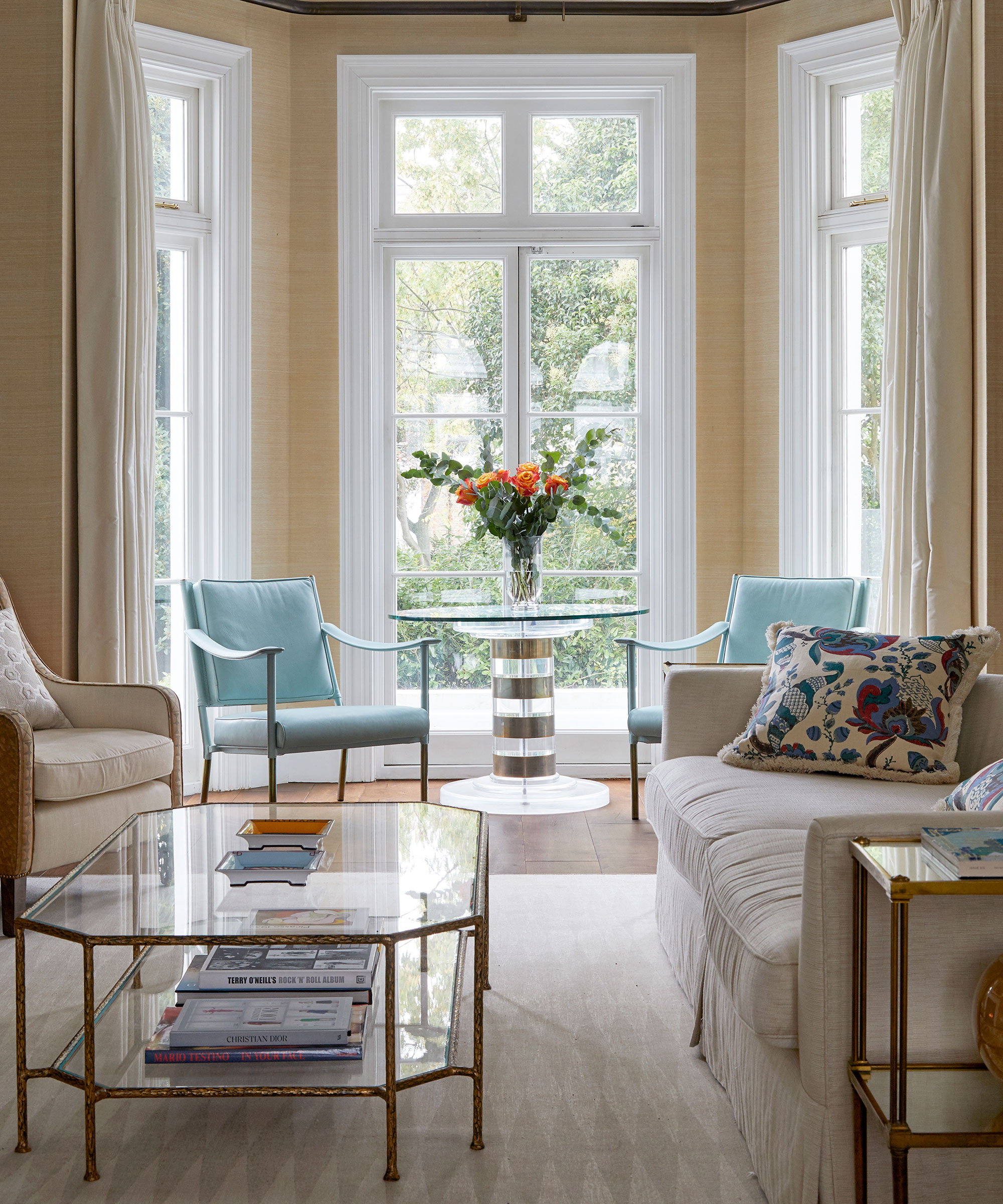 An example of beige living room ideas with textured wallpaper, blue accent chairs, a glass coffee table and large bay windows.