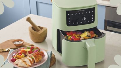 Pastel blue kitchen with the Aldi pastel green air fryer with cooked vegetables in the tray next to a plate of chicken and veg on the countertop