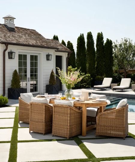 a poolside wicker and wood dining table styled on a diamond patio with a large white poolhouse in the background
