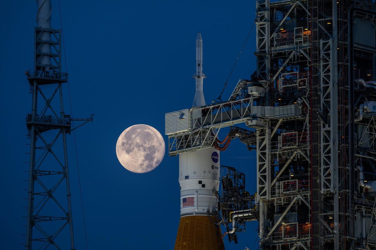 the full moon hangs in frame with the orion, inside its payload shell atop the SLS rocket.