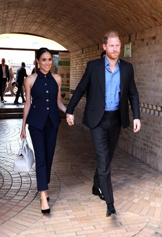 Meghan Markle wears a white Loro Piana handbag with navy pants, a navy vest and black pumps.