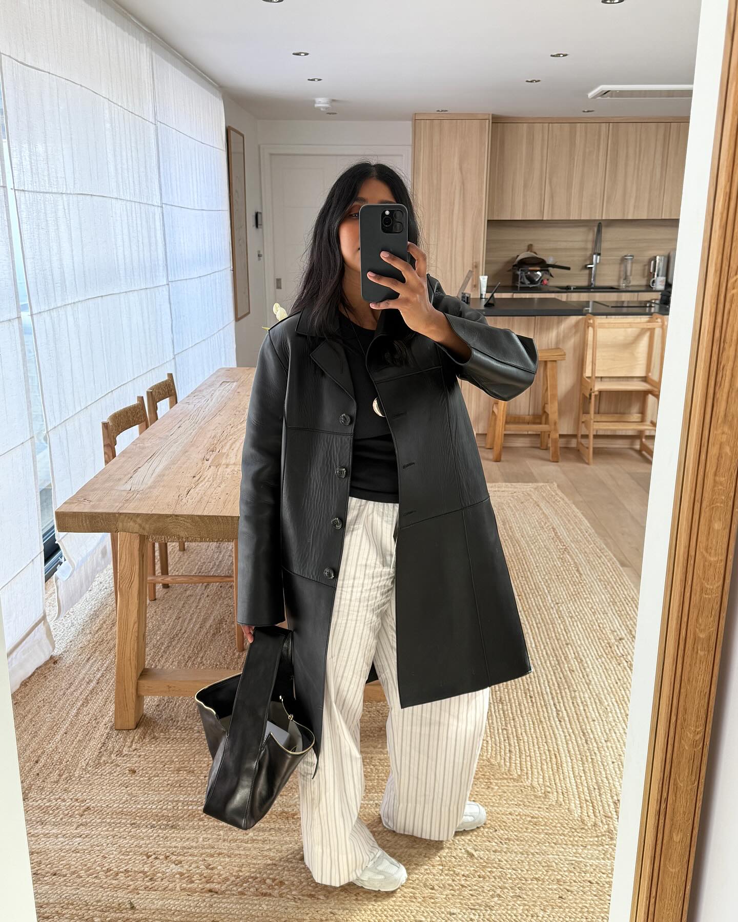 An image of @monikh wearing a leather trench coat, striped linen trousers and white trainers, an example of an autumn trainer outfit.