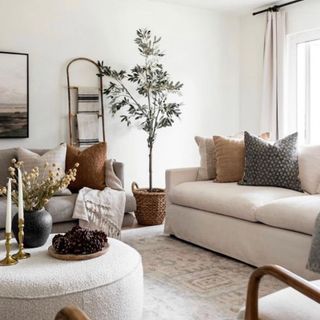Warm neutrals scheme in lounge with round boucle ottoman coffee table, large olive tree in basket, rustic towel ladder, and tonal scatter pillows on comfy sofas.