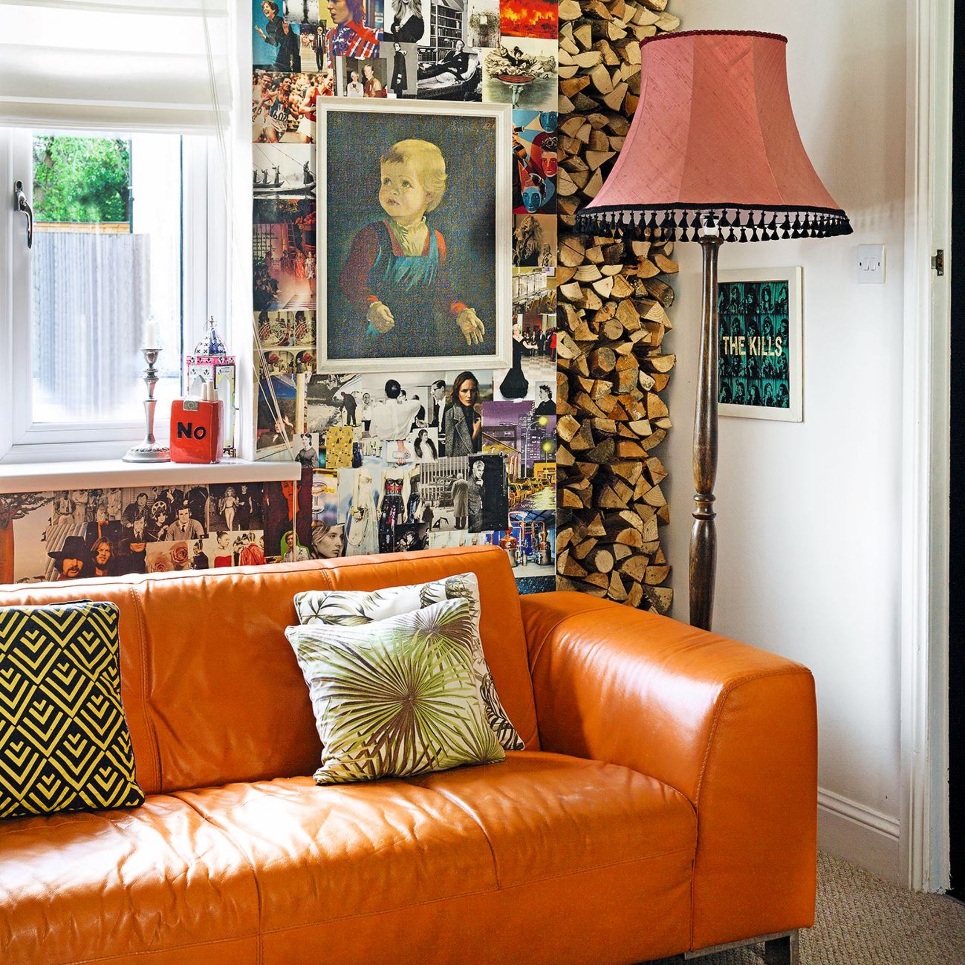 Living room detail with bright orange leather sofa on beige carpet, vintage floor lamp with red shade and tassels, firewood stacked in niche and wall covered in magazine cuttings