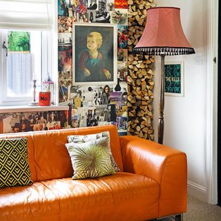 Living room detail with bright orange leather sofa on beige carpet, vintage floor lamp with red shade and tassels, firewood stacked in niche and wall covered in magazine cuttings
