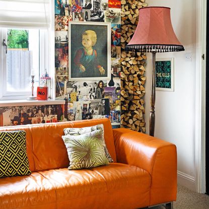 Living room detail with bright orange leather sofa on beige carpet, vintage floor lamp with red shade and tassels, firewood stacked in niche and wall covered in magazine cuttings