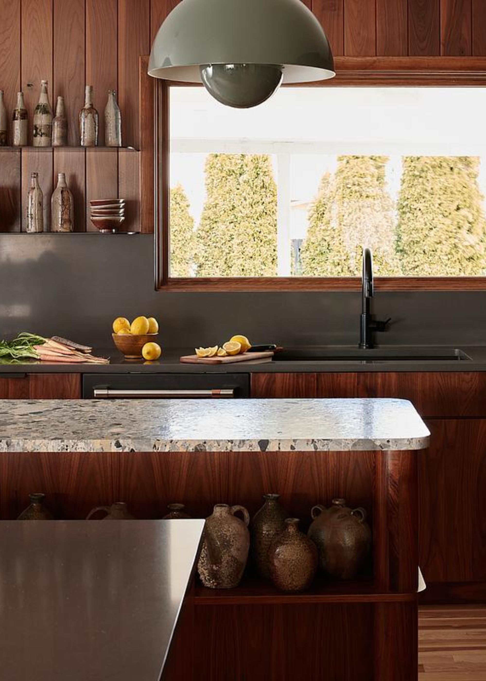A dark wood kitchen with an island in the middle and a large sage pendant light hanging above. There is also a large window looking into the garden and double open shelving units beside the window with kitchen essentials on top. On the countertop is a bowl of lemons and sliced up lemons as well as a batch of uncut carrots.