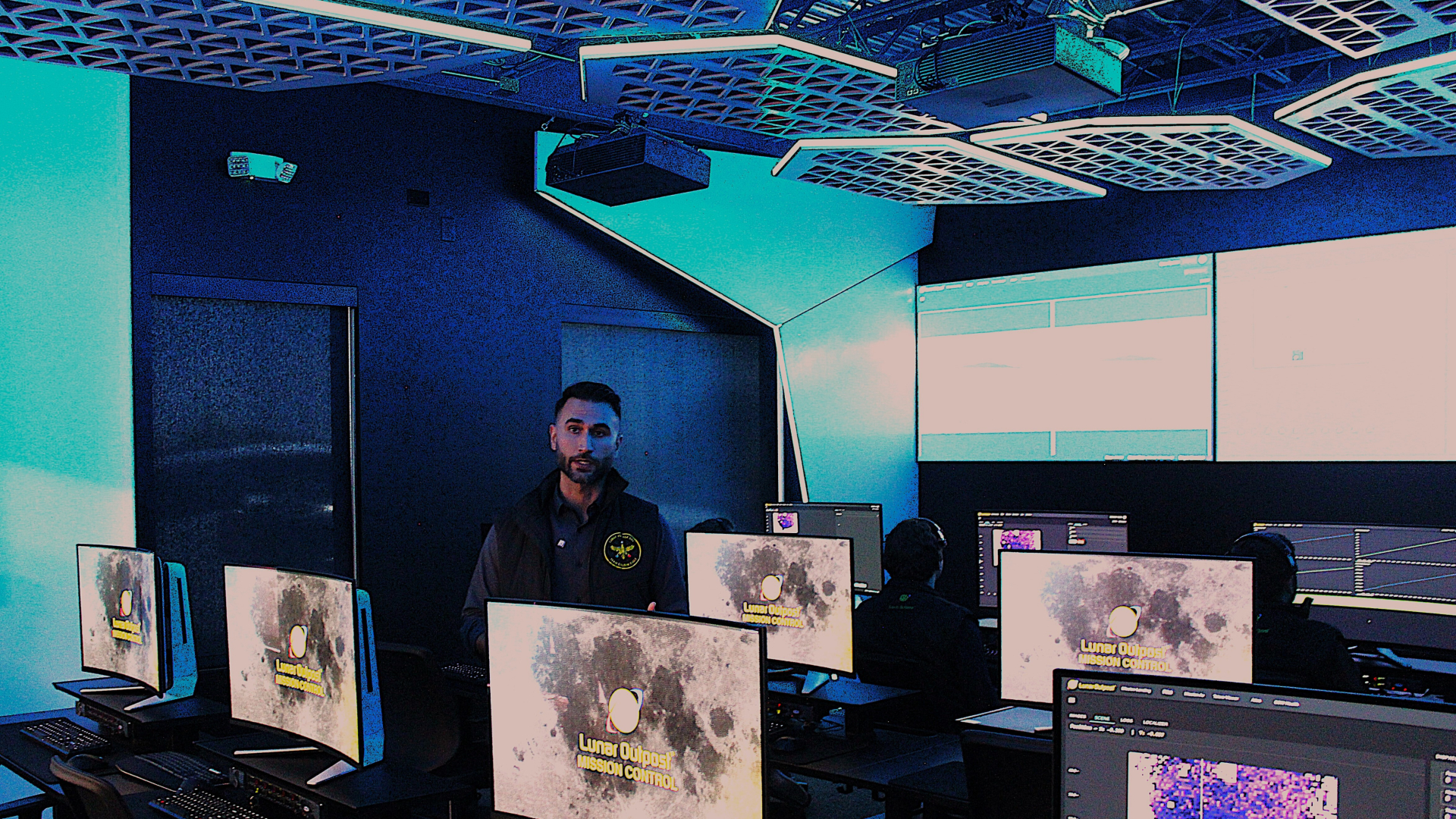 A man stands behind a row of computers in a dimly lit room