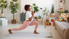 Woman practicing yoga in living room. She is in low lunge pose, with her front leg bent at the knee and foot flat on the floor, and her rear leg extended behind and on her toes. She holds her palms together in front of her chest. She is wearing pink leggings and a white T-shirt.