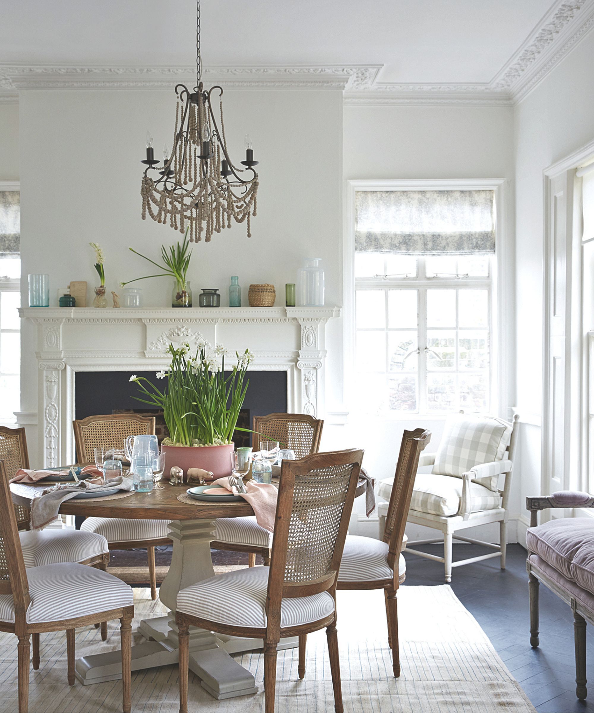 Rustic dining room with a large fireplace, a round wooden table, rattan dining chairs, a chandelier above, and a large potted narcissus in the middle.
