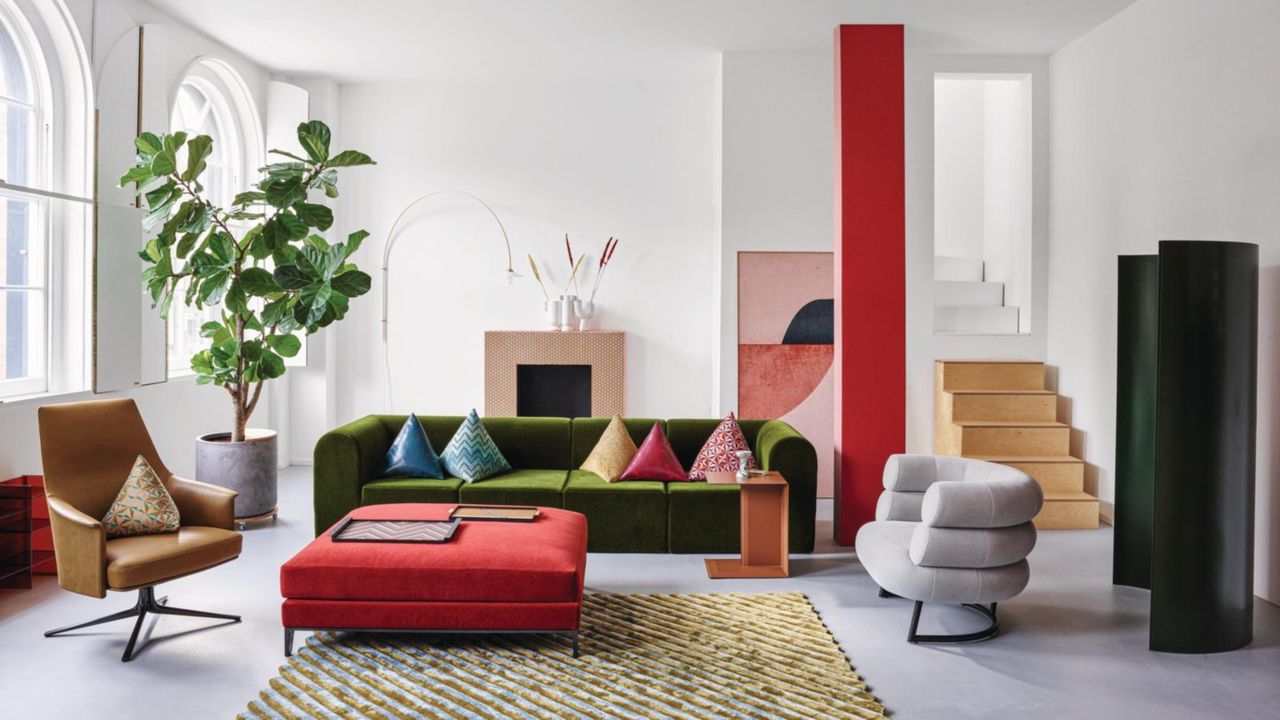 A large, white living room with colorful pops. There is an olive green velvet couch, a bright red-painted beam, a red square ottoman and a large green potted plant near the window