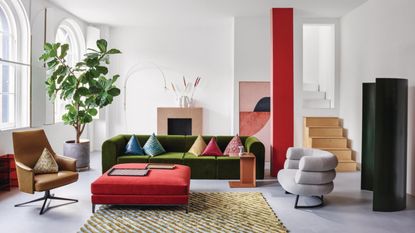 A large, white living room with colorful pops. There is an olive green velvet couch, a bright red-painted beam, a red square ottoman and a large green potted plant near the window