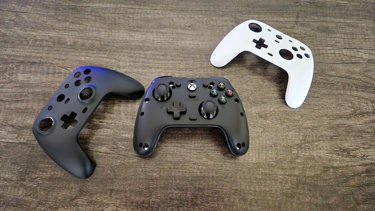 Two gaming controller shells are arranged on a wooden surface next to an exposed Xbox controller. One shell and the controller are black, and one shell is white, showcasing a sleek, minimalist design.