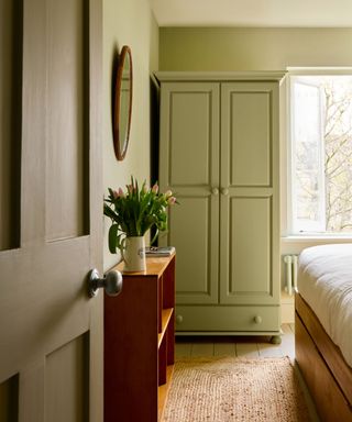 Green bedroom with light green wardrobe, wooden console with vase of tulips and woven rug