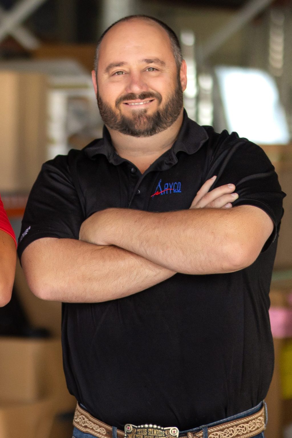 Caleb Caviness with arms crossed, in black polo shirt, in front of blurred-out warehouse.