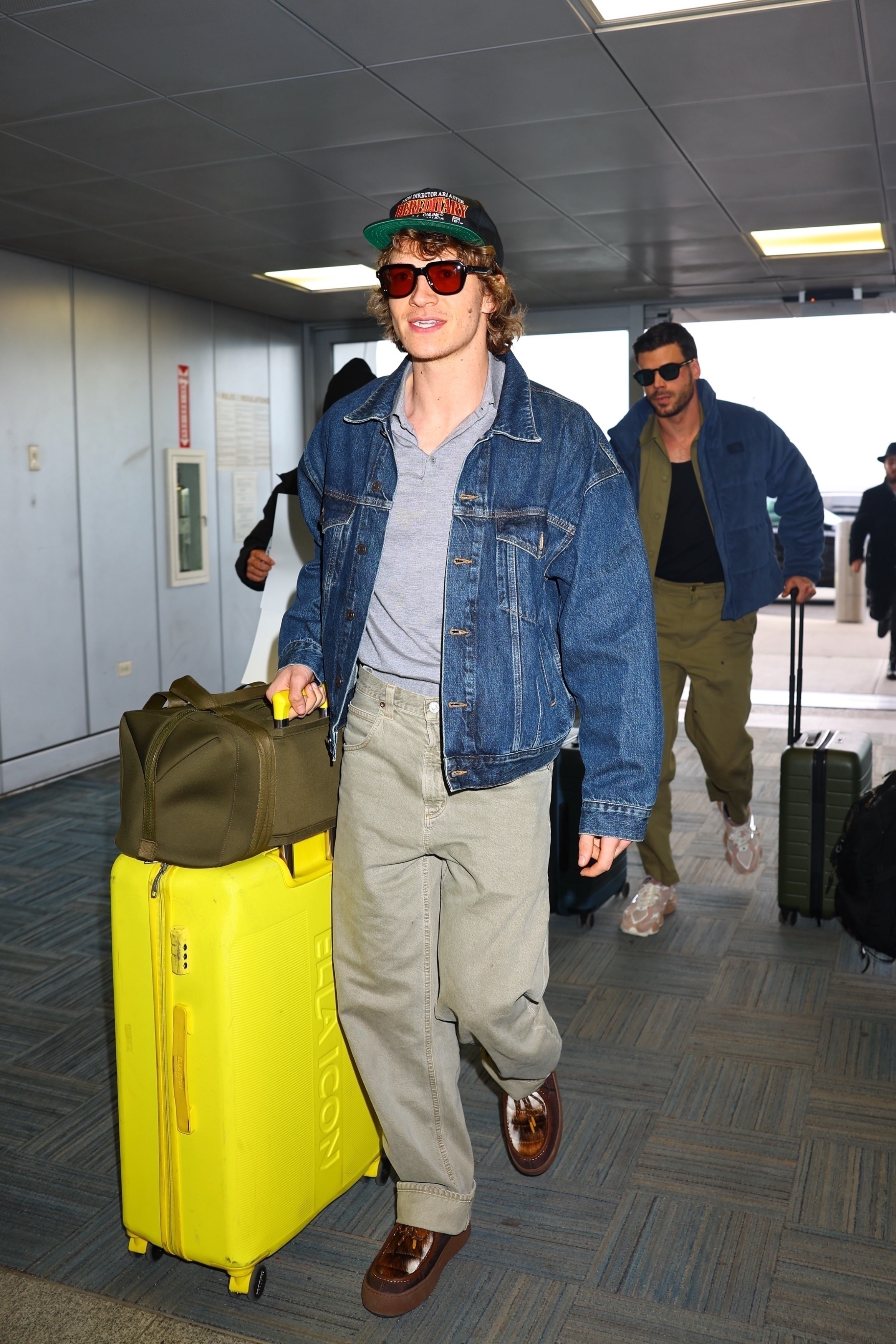 Connor wears a denim jacket, gray sweater, gray-washed jeans, brown shoes, a dagne dover green bag, and yellow suitcase.