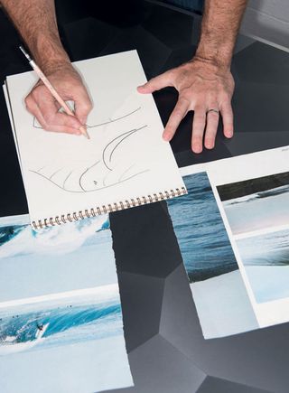 View of Alex Rasmussen's hands creating a sketch. There are images of waves on the table in front of the sketch pad
