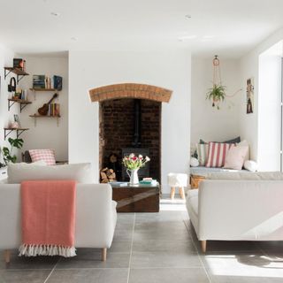 Open living room with white walls, grey stone floor, off-white corner sofas, wooden panel, brick fireplace, and colourful decorative accents A renovated Victorian four bedroomed farmhouse in Ottery St Mary, Devon, home of Emma and Hendrick Jaulin