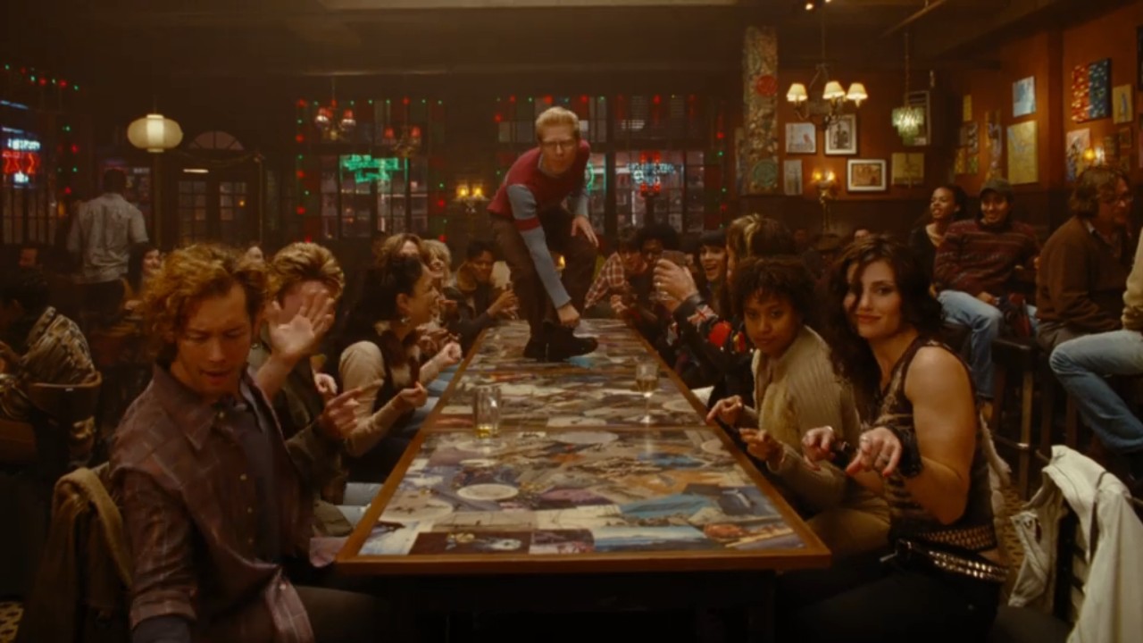 The cast of rent sitting at a table while Mark stands on it.