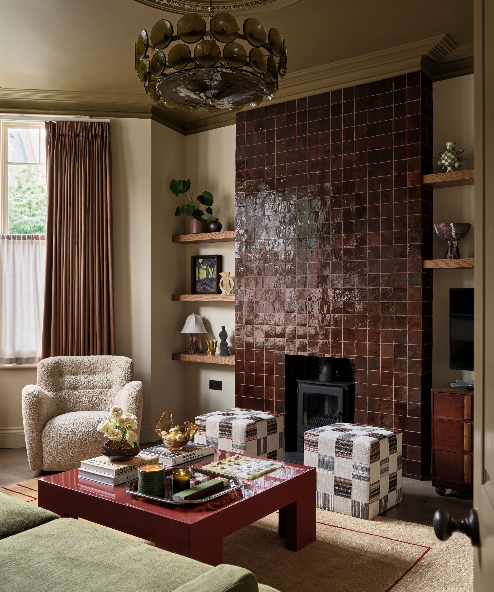 Living room with maroon tiled feature wall, log-burner, a red lacquer coffee table with decor on top, two square ottomans, a boucle armchair and a green velvet sofa, and shelves beside fireplace