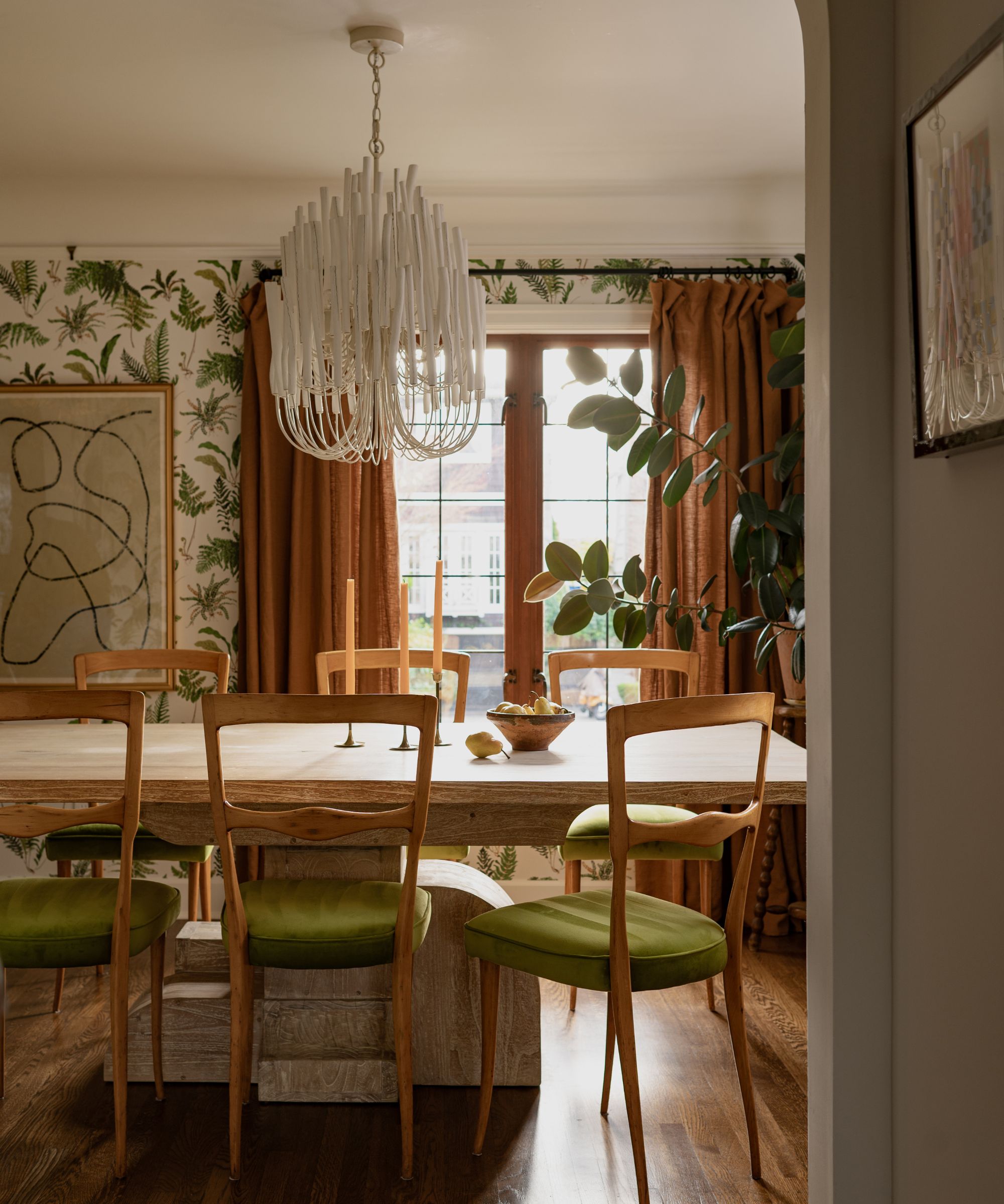Dining room with green and cream botanical wallpaper, large wooden dining table with wooden chair upholstered in green velvet and large potted plant in one corner