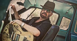 Dave Stewart at the wheel of a pickup truck wearing a hat and shades, and holding his Duesenberg semi-hollow.