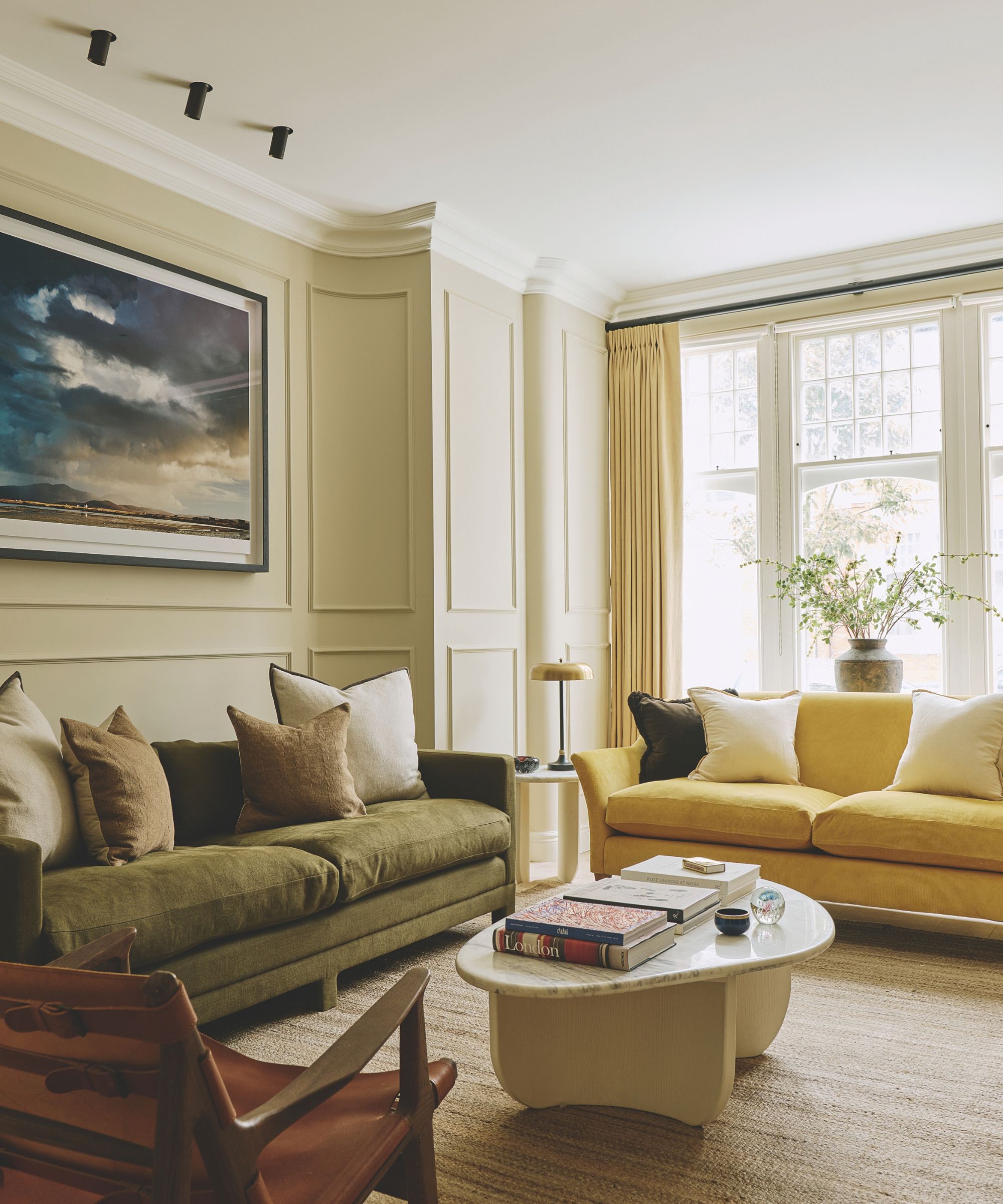 Elegant living room with paneled cream walls, olive green velvet sofa, yellow accent couch, marble coffee table, and large window with curtains
