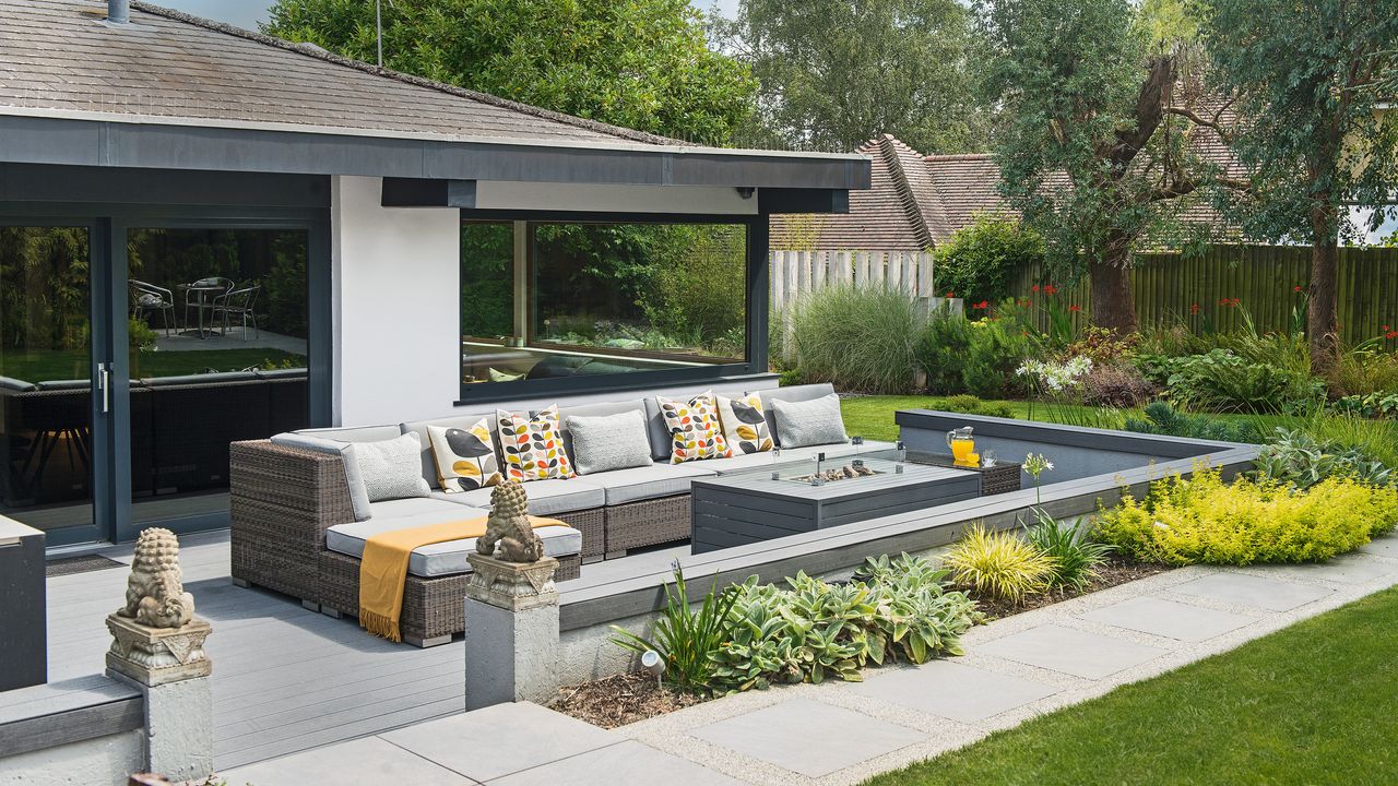 a decked area of a garden with a large corner outdoor sofa with cushions and a firepit table