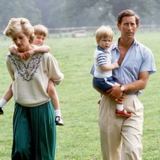 A 1980s photo of Princess Diana wearing a long green skirt and white sweater giving Prince William a piggyback in grass next to Prince Charles, who is wearing khakis and a blue shirt holding Prince Harry