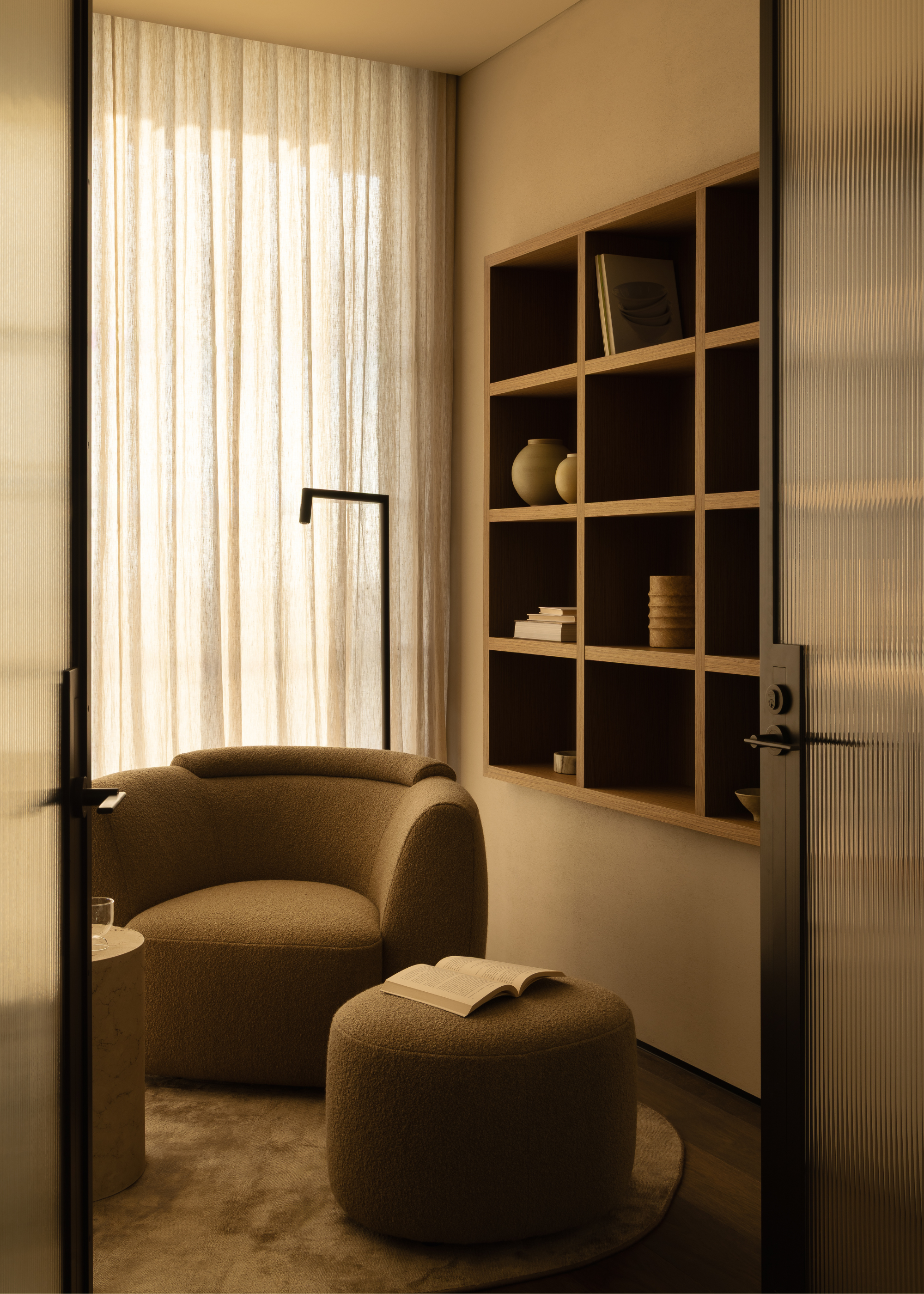 A study space with in-built storage shelves, a round suede rug, a plush armchair and a pouff by a floor lamp and large windows with sheer treatment