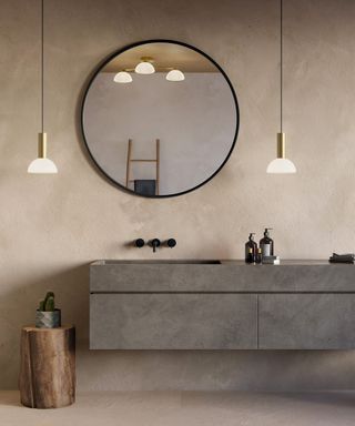 A grey bathroom sink unit with large round mirror and twp black pendant lights hanging. In the reflection a flush ceiling light with three bulbs
