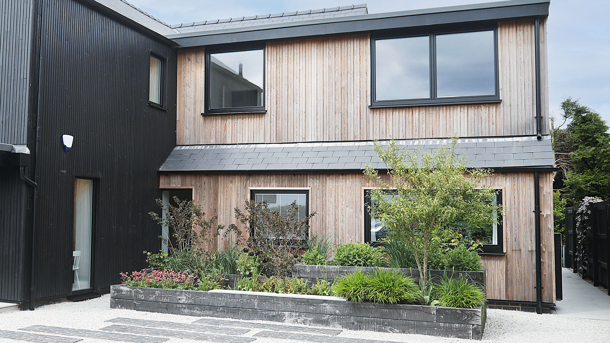 timber-clad house with a gravel driveway and small front garden area with raised beds, shrubs and a small tree