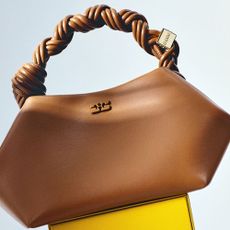 Brown Ganni bag with a twisting handle on a yellow box and white background