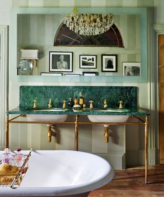 Green bathroom with green striped painted walls, large mirror above green marble vanity unit with brass detailing, warming wooden floor, corner of white bathtub