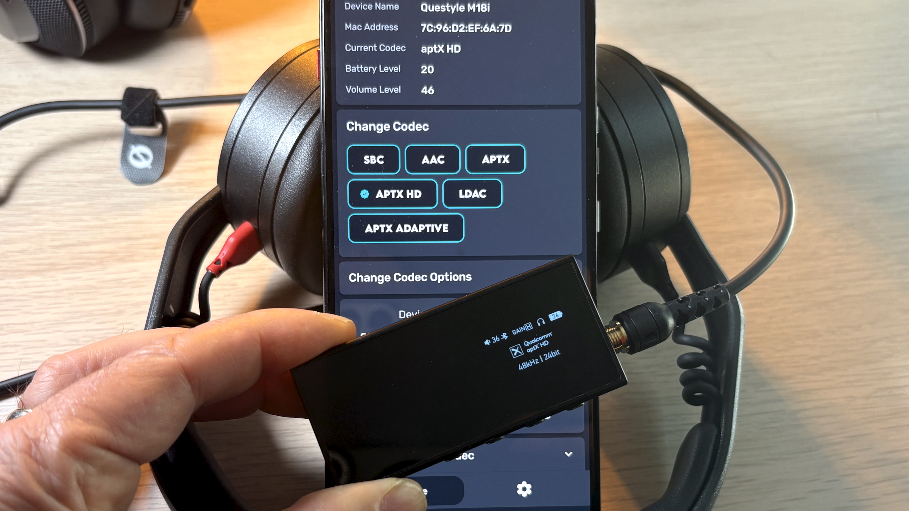 A hand holding a DAC, in front of a phone screen showing the Bluetooth codec changer app
