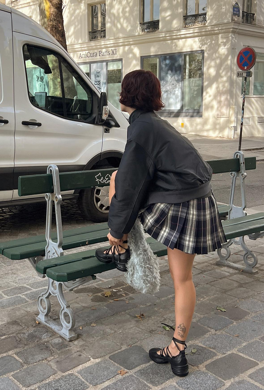 The best ballet sneakers are shown in a photo of a woman standing outside near a bench in Paris wearing a black leather bomber jacket with a navy plaid pleated mini skirt and black lace-up ballerina sneakers