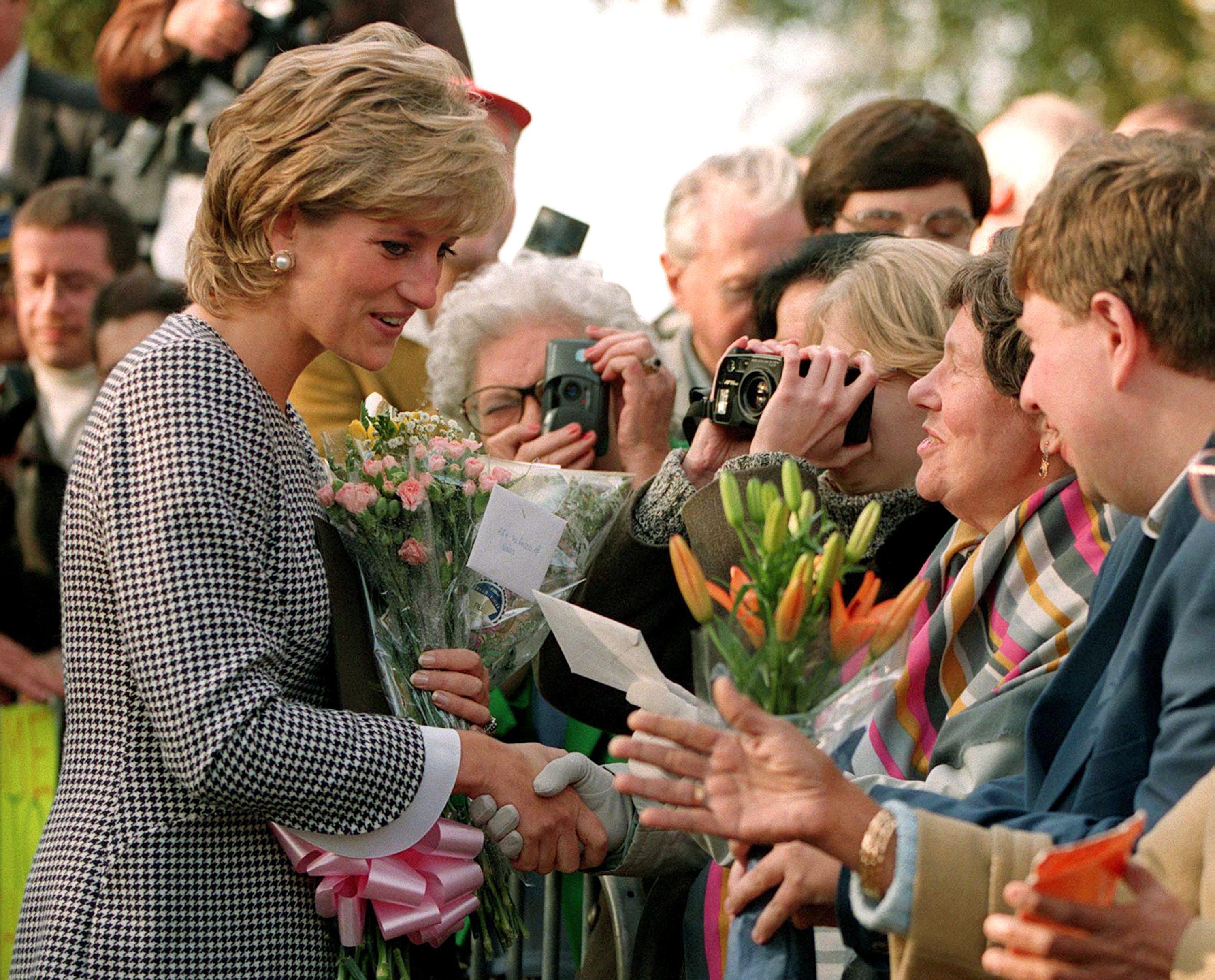Princess Diana wearing a black houndstooth coat talking to a crowd of fans holding flowers